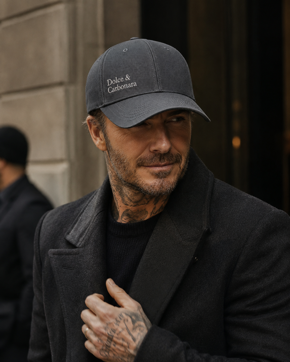 A close-up portrait of David Beckham wearing a dark gray baseball cap with the text 'Dolce & Carbonara' on the front and a matching dark coat.