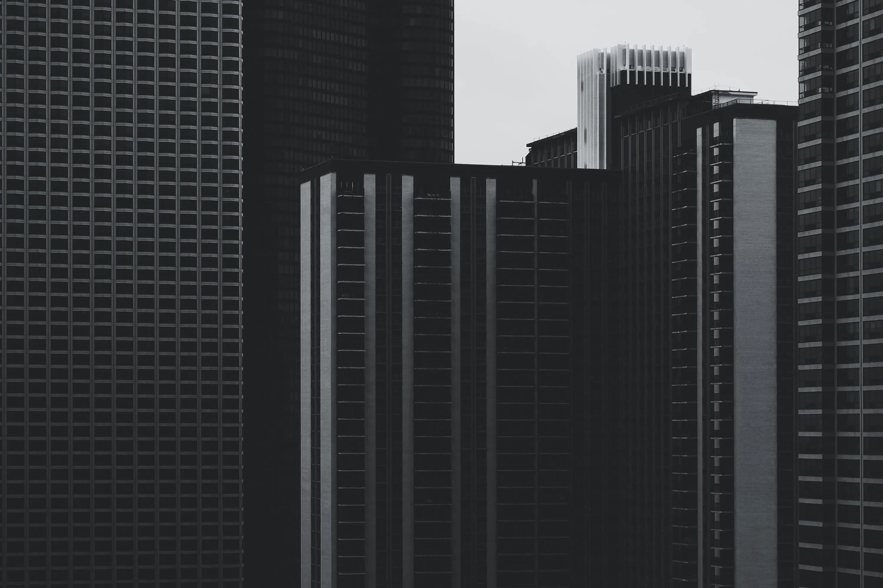 A monochromatic low-angle shot of several modern skyscrapers with repeating geometric architectural patterns. The high-contrast composition emphasizes the vertical lines and grid-like facade of the buildings against a muted sky.
