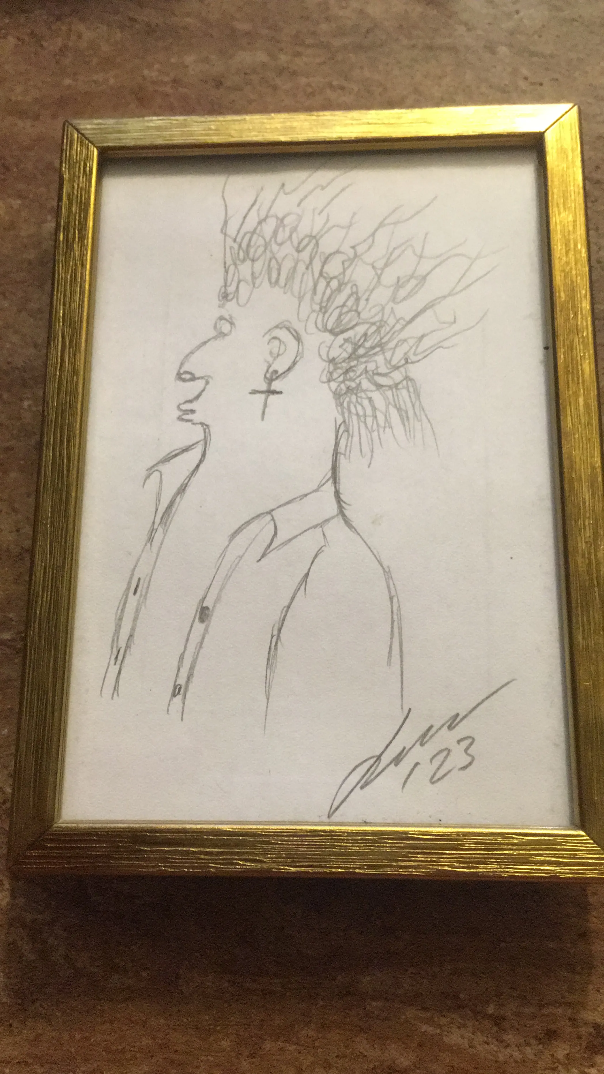 A pencil sketch in profile of a person with wild, scribbled hair and a cross earring, housed in a gold frame. The drawing includes a signature and the year 23 in the lower right corner.
